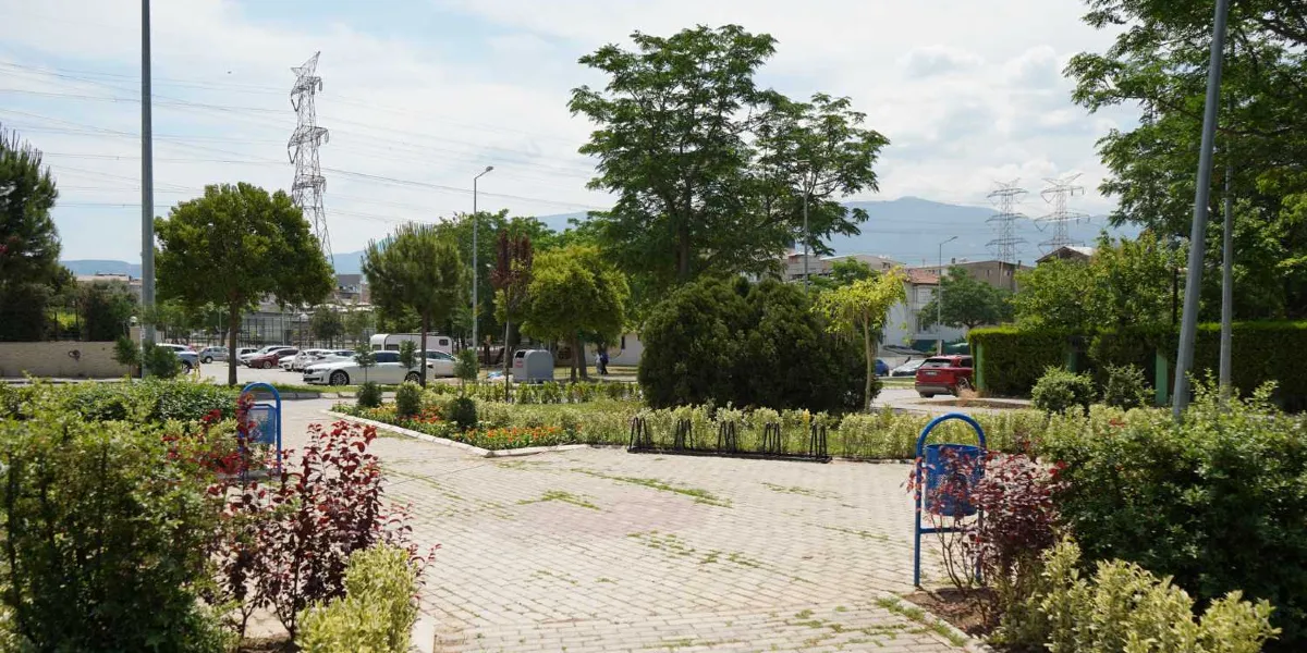 Ağaçlarla çevrili, parke taşlı yürüyüş yolları, yeşil alanları ve mavi çöp kutuları bulunan park alanı. Arka planda elektrik iletim hatları, ağaçlar ve park halindeki araçlar görülüyor.