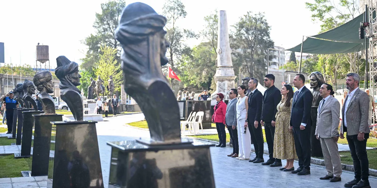 Bornova’da bir parkta, yan yana dizilmiş tarihi figürlerin büstlerine karşı saygı duruşunda bulunan bir grup resmi giyimli kişi. Arka planda yüksek bir anıt ve Türk bayrağı görülüyor.