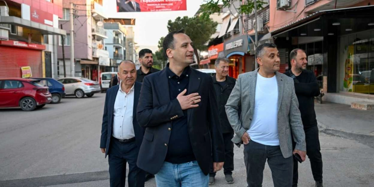 Egemenlik Mahallesi'ndeki bir sokakta yürüyen Başkan Eşki, lacivert ceketli ve kot pantolonlu haliyle sağ elini kalbine koyarak selam veriyor. Başkan'a, aralarında gri ceketli ve beyaz tişörtlü bir kişinin de bulunduğu bir grup erkek eşlik ediyor. Arka planda dükkanların sıralandığı cadde ve park halindeki araçlar görülüyor, üst kısımda ise kırmızı bir teşekkür pankartı asılı duruyor.