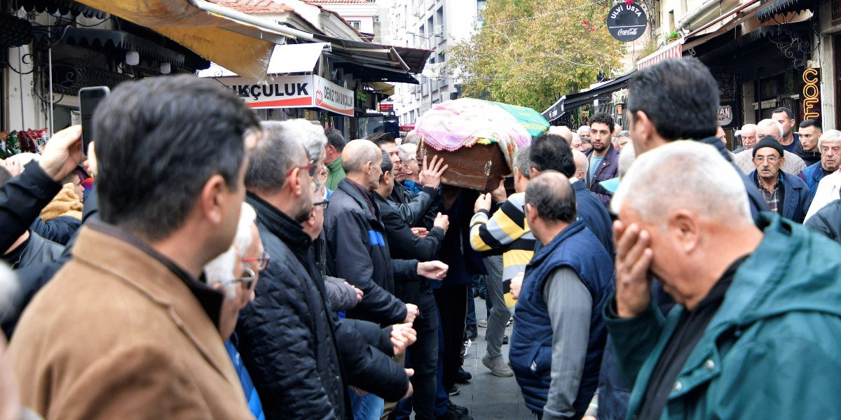 Kalabalık bir grup insanın, omuzlarında taşıdıkları bir tabutu Bornova'daki bir çarşı içinden geçirdiği cenaze töreni. İnsanların birçoğu üzgün bir ifadeyle tabutu takip ederken, ön planda yüzünü elleriyle kapatan yaslı bir adam dikkat çekiyor.