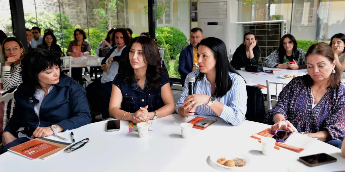 Aydınlık bir iç mekanda, yuvarlak bir masanın etrafında toplanmış bir grup kadın ve erkek bir toplantıya katılıyor. Ön planda, masanın merkezinde oturan kadınlardan biri elinde mikrofon tutarak konuşma yapıyor; yanındaki diğer kadınlar ise onu dikkatle dinliyor. Masanın üzerinde akıllı telefonlar, kağıtlar ve üzerinde 