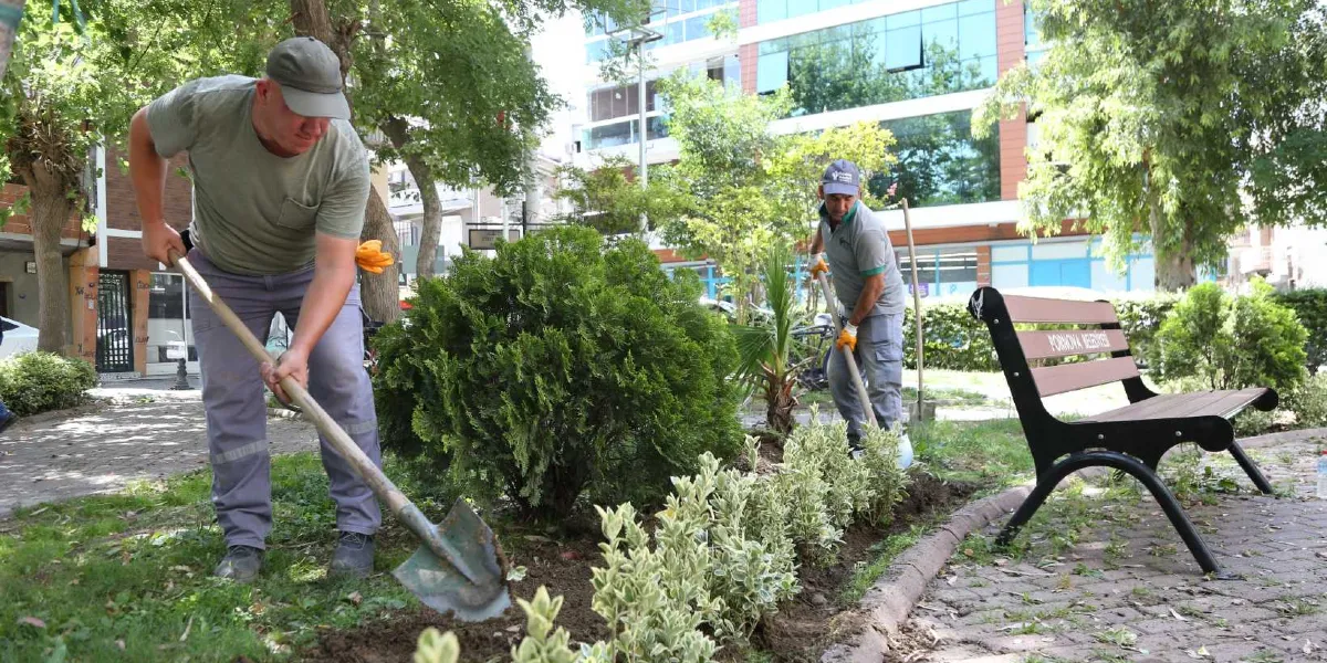 Bornova Belediyesi personeli, park alanında ellerinde küreklerle yeni fidan dikimi ve çevre düzenlemesi yapıyor. Ön planda, üzerinde Bornova Belediyesi yazılı ahşap bir bankın bulunduğu yeşil alanda çalışan iki işçi görülüyor.