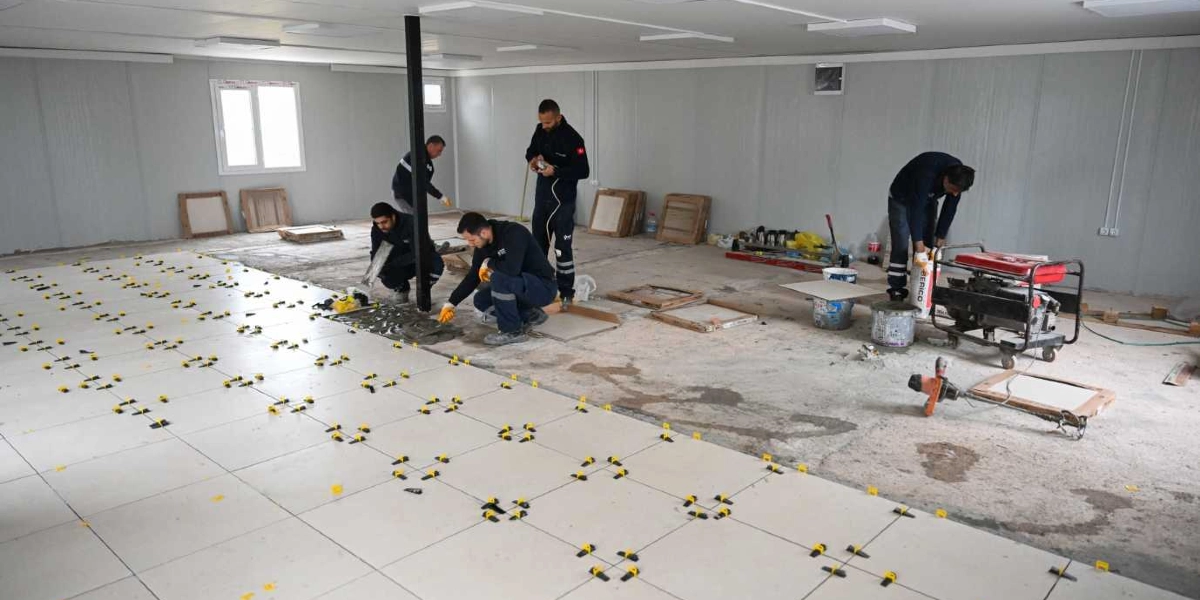 İnşaat halindeki geniş bir kapalı alanda, iş kıyafetleri giymiş dört işçi zemin döşeme çalışması yapıyor. İşçilerden ikisi diz çökmüş vaziyette yer karolarını yerleştirirken, diğerleri harç hazırlığı ve malzeme taşıma işlemleriyle ilgileniyor. Zeminin bir kısmına beyaz fayanslar döşenmiş ve aralarına hizalama için sarı plastik klipsler yerleştirilmiş, alanın geri kalanı ise henüz beton zemin halindedir.
