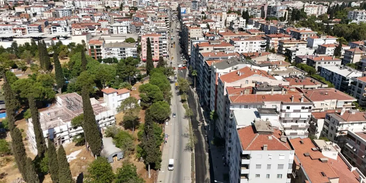 Bornova'da kuş bakışı çekilmiş, yoğun yerleşimli binaların ve ağaçlık alanların arasında uzanan asfaltlanmış bir ana cadde. Yolun bir şeridi yeni dökülmüş koyu renkli asfaltla kaplıyken, diğer şeridinde araç trafiği akmaya devam ediyor.