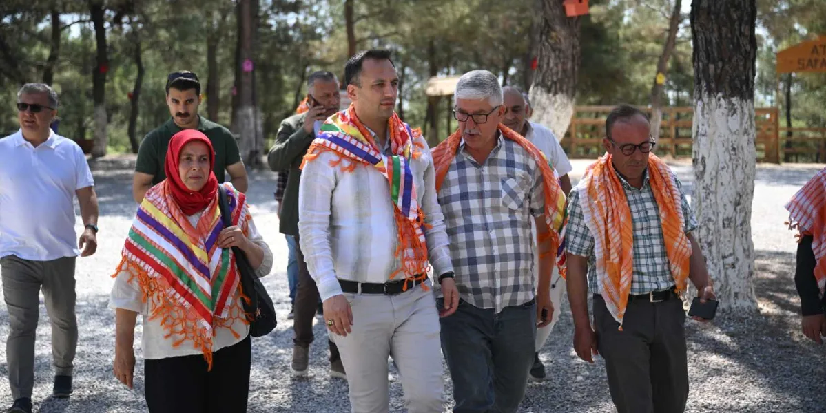 Ağaçlık bir alanda yürüyen, omuzlarında renkli yöresel şallar bulunan bir grup insan. Önde, kırmızı başörtülü bir kadın, yanında açık renkli gömlekli bir erkek ve onun yanında kareli gömlekli, gözlüklü bir erkek yer alıyor. Arka planda ise farklı kıyafetler giymiş diğer katılımcılar ve ağaç gövdelerine asılmış kuş evleri görülüyor.