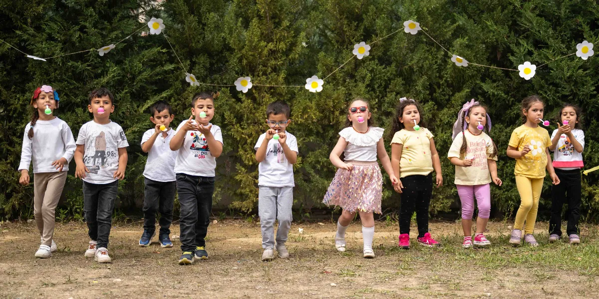 Yeşil ağaçların önünde yan yana dizilmiş, ağızlarında renkli lolipoplar tutan dokuz çocuk, Anneler Günü kutlaması kapsamında bir yarışma veya etkinlik için poz veriyor. Çocukların arkasında, ipe dizilmiş beyaz papatya süslemeleri bulunuyor. Çocukların bir kısmı beyaz tişörtler, bir kısmı ise sarı ve renkli kıyafetler giymiş durumda; aralarından biri güneş gözlüğü takıyor, bir diğeri ise saçında çiçekli bir taç taşıyor.