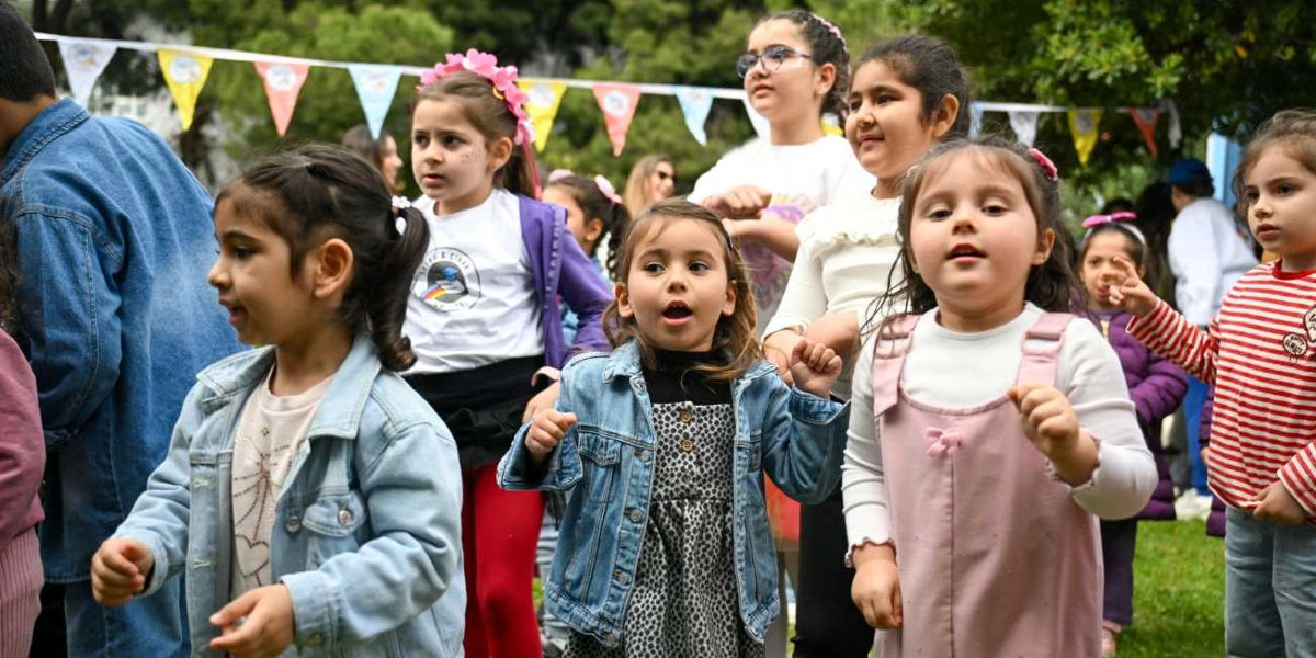 Açık havada, üzerinde renkli üçgen flamaların asılı olduğu bir alanda dans eden veya eğlenen bir grup çocuk. Ön planda, kot ceket giymiş küçük bir kız çocuğu, yanında pembe tulumlu bir başka çocuk ve arka planda beyaz tişörtlü, saçında çiçekli taç olan bir kız çocuğu ile diğer çocuklar neşeli bir şekilde yer alıyor. Çocukların çoğu günlük kıyafetler içinde ve ortamda şenlik havası hakim.