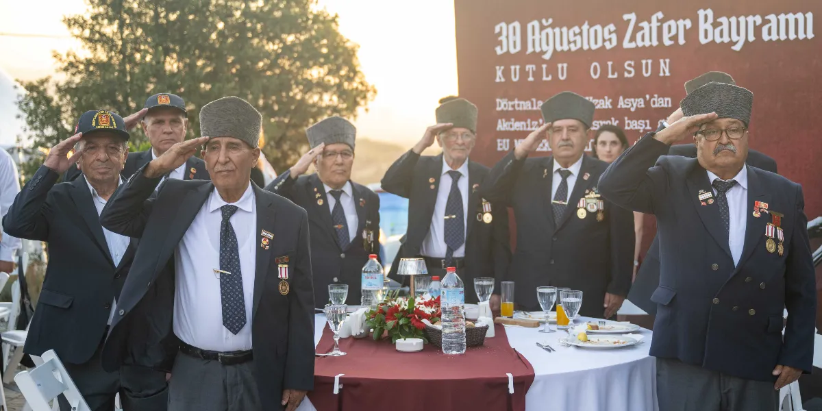 Koyu renkli takım elbiseleri, yakalarında madalyaları ve başlarında kalpaklarıyla bir masanın etrafında duran gaziler, asker selamı veriyor. Arka planda üzerinde 