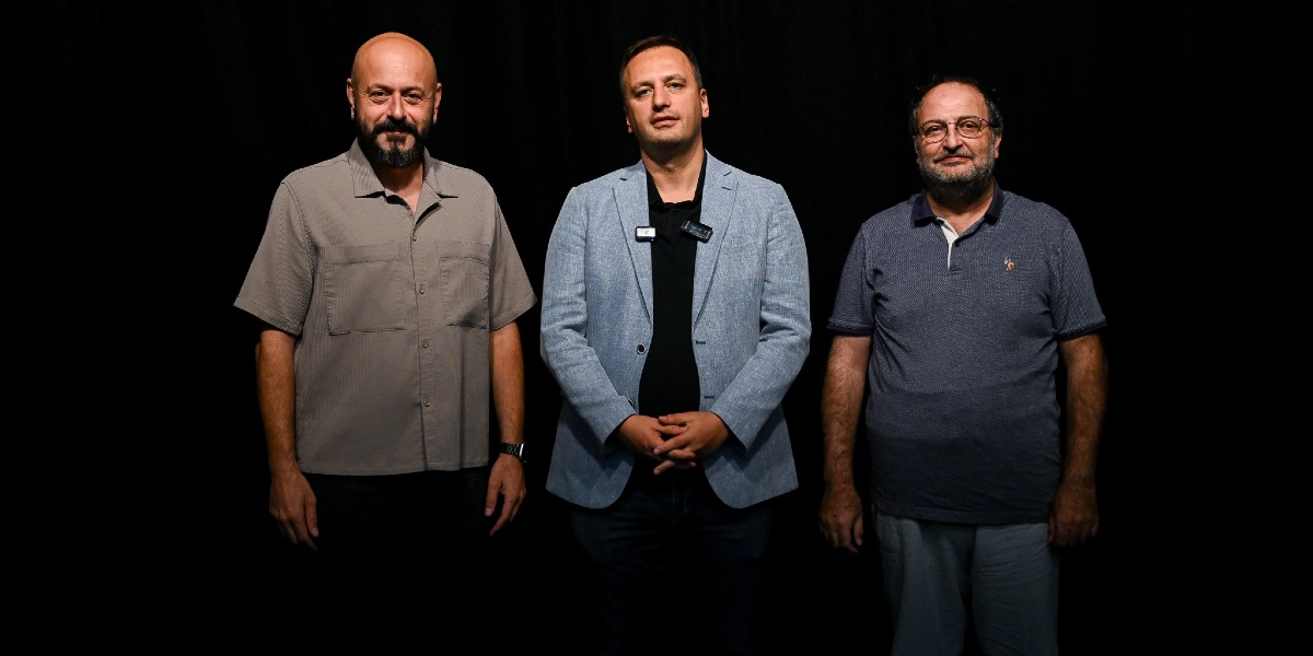 Siyah bir arka plan önünde yan yana duran üç erkek. Soldaki kişi kısa saçlı, sakallı, bej rengi kısa kollu bir gömlek giyiyor. Ortadaki kişi kısa saçlı, siyah tişört üzerine açık mavi bir ceket giymiş ve yakasında bir yaka mikrofonu bulunuyor. Sağdaki kişi ise gözlüklü, sakallı ve lacivert polo yaka bir tişört giyiyor.