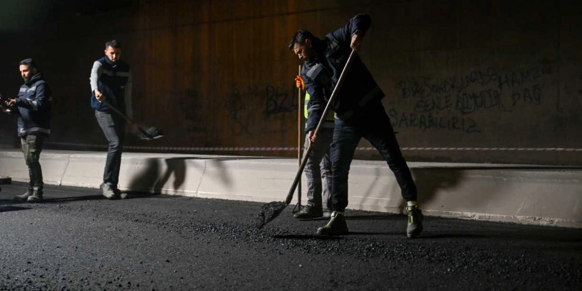Gece vakti bir tünel içerisinde, iş kıyafetleri giymiş üç işçi ellerindeki küreklerle yeni dökülen asfaltı düzeltiyor. İşçilerden biri ön planda eğilmiş halde küreğiyle asfaltı yayarken, diğer ikisi arka planda çalışmaya devam ediyor. Arka taraftaki beton duvarda sprey boya ile yazılmış yazılar ve grafitiler bulunuyor.