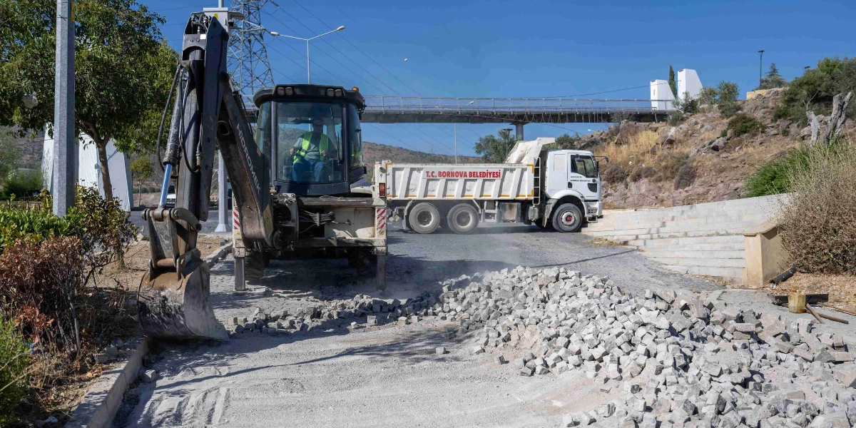 Hidromek marka bir iş makinesi, üzerinde T.C. Bornova Belediyesi yazan beyaz bir damperli kamyonun yanında, sökülmüş parke taşlarının bulunduğu bir yol çalışması alanında görev yapıyor. Operatör koltuğunda sarı yelekli bir çalışan oturuyor ve arka planda bir yaya üst geçidi ile kayalık bir yamaç görülüyor.