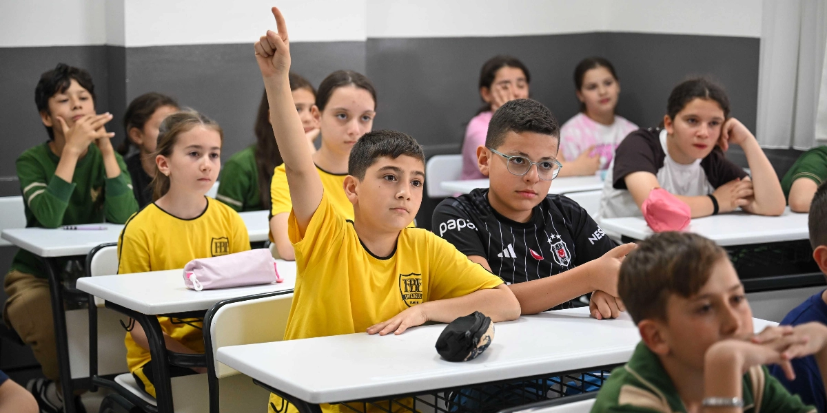 Sınıf ortamında sıralarda oturan ortaokul çağındaki öğrenciler, dikkatle dersi dinliyor. Ön planda sarı forma giyen bir erkek öğrenci söz almak için elini kaldırmış, yanında ise siyah forması ve gözlükleriyle bir başka öğrenci oturuyor. Diğer öğrenciler de sıralarında oturarak dersi takip ediyor.