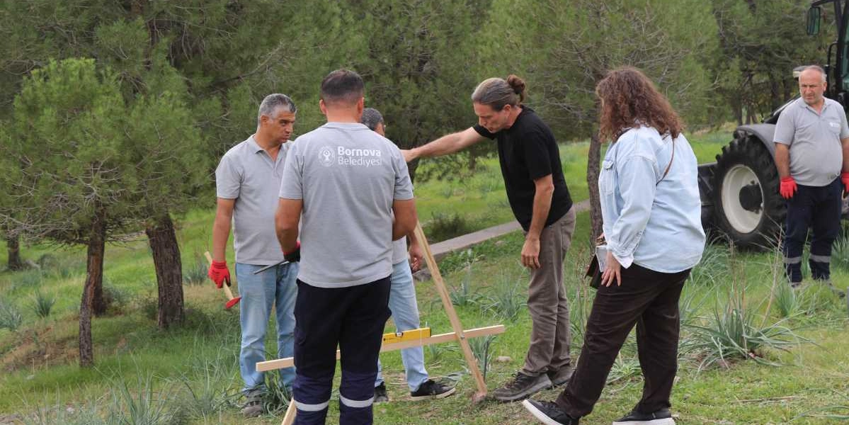 Açık havada, ağaçlık bir alanda Bornova Belediyesi logolu tişört giyen görevlilerin de aralarında bulunduğu bir grup insan, arazi üzerinde ahşap bir ölçüm düzeneğiyle inceleme yapıyor. Grubun yanında bir traktör bulunurken, ekip üyeleri doğa temelli çözümler üzerine çalışma yürütüyor.