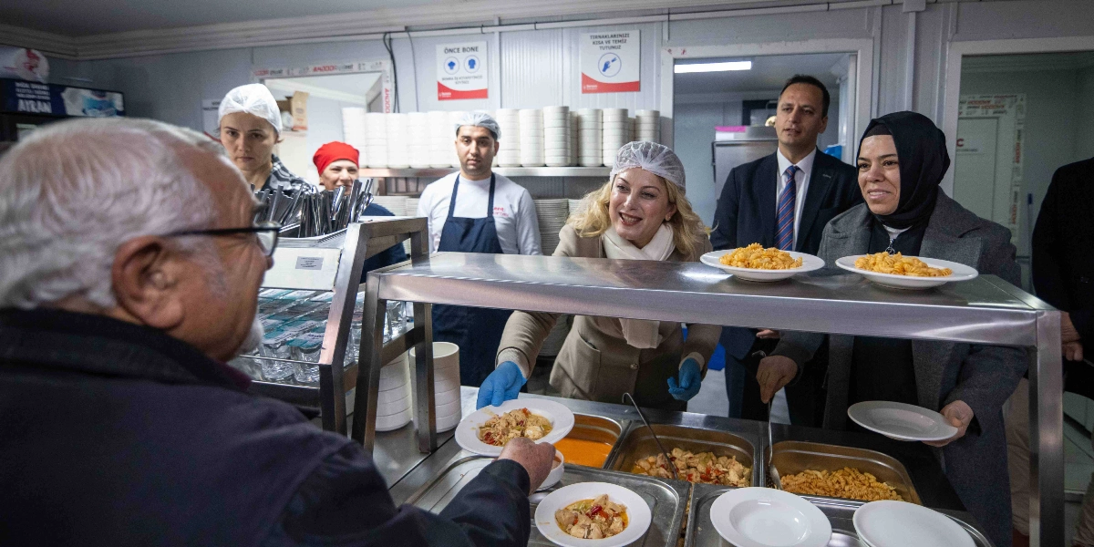Bir aşevi veya yemekhanede, servis tezgahının arkasında duran CHP Kadın Kolları Başkanı Asu Kaya ve beraberindeki heyet, yaşlı bir vatandaşa yemek ikram ediyor. Asu Kaya, başında bone ve ellerinde mavi eldivenlerle tabaklara yemek koyarken gülümsüyor. Yanında başörtülü bir kadın ve takım elbiseli bir erkek yer alıyor. Arka planda ise mutfak personeli ve duvarda hijyen kurallarını hatırlatan uyarı levhaları görülüyor.