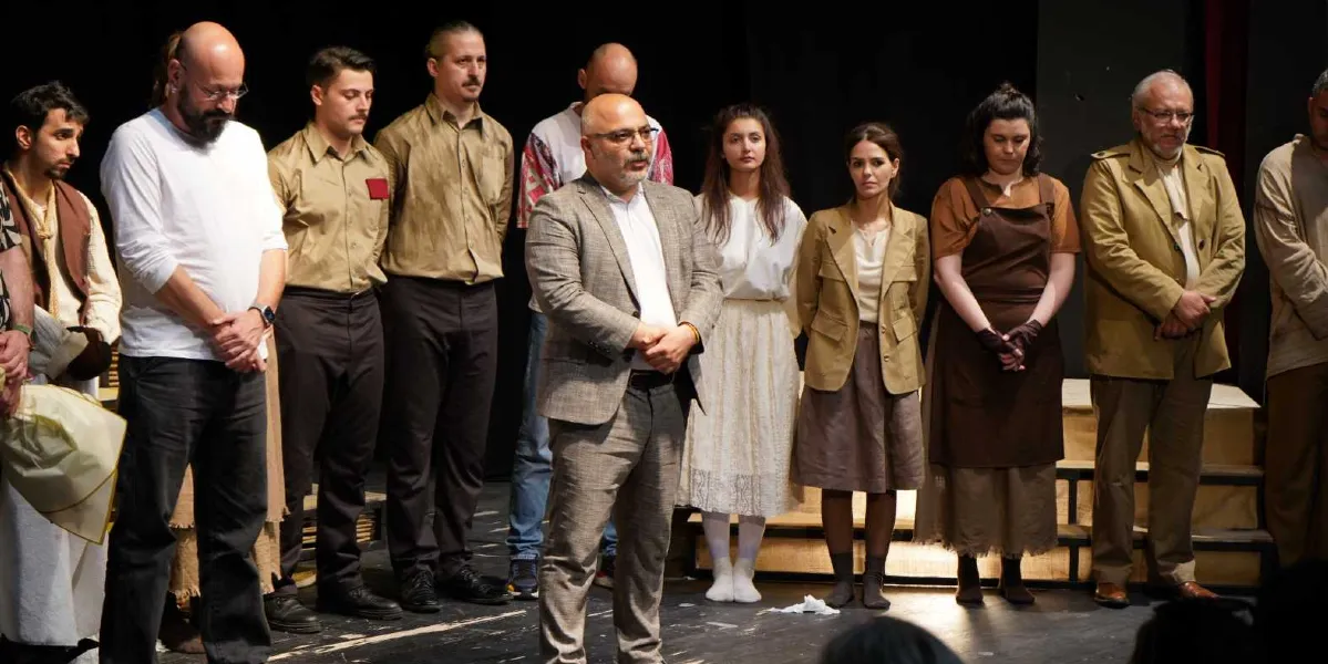 Tiyatro sahnesinde yan yana dizilmiş, farklı dönem kıyafetleri giyen bir grup oyuncu ve merkezde konuşma yapan gri ceketli bir adam. Oyuncular, beyaz gömlekler, kahverengi tonlarında ceketler, etekler ve önlükler gibi çeşitli kostümler içinde, elleri önlerinde birleşmiş şekilde duruyorlar. Sahne ışıkları altında, seyircilere dönük bir selamlaşma veya kapanış anı sergileniyor.