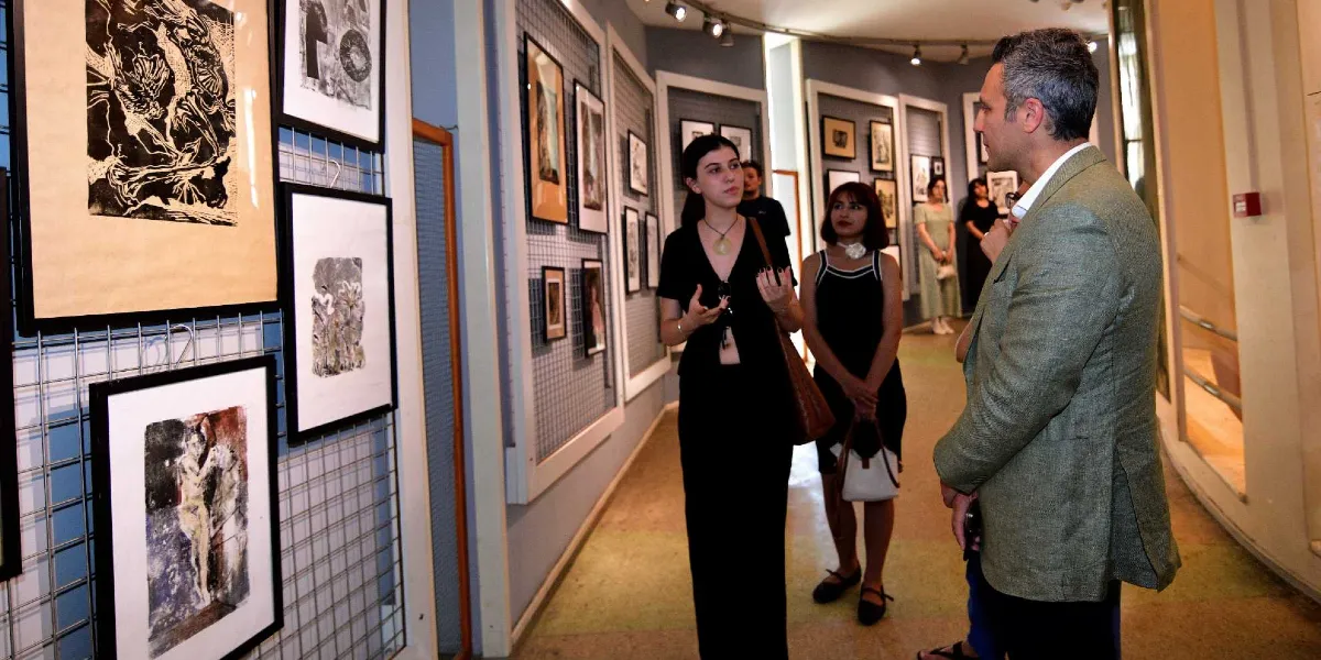 Bornova'daki bir sanat galerisinde, duvarlardaki ızgara panellere asılı siyah beyaz baskı eserleri inceleyen bir grup insan. Ön planda, siyah kıyafetli genç bir kadın, yanında duran diğer kişilere eserler hakkında bilgi veriyor; karşısında ise yeşil ceketli bir adam onu dinliyor. Arka planda ise sergiyi gezen diğer ziyaretçiler görülüyor.