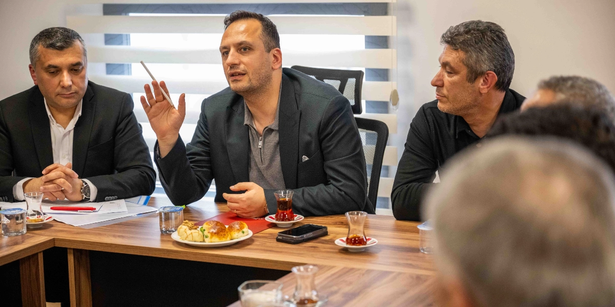 Modern bir ofis ortamında, ahşap bir toplantı masası etrafında bir araya gelmiş erkeklerden oluşan bir grup. Masanın ortasında oturan ve elinde bir kalemle konuşan kişi, diğer katılımcılara hitap ediyor. Masanın üzerinde ince belli çay bardakları, bir tabak poğaça ve bir cep telefonu bulunuyor. Katılımcılar resmi ve yarı resmi kıyafetlerle toplantıya katılıyor.