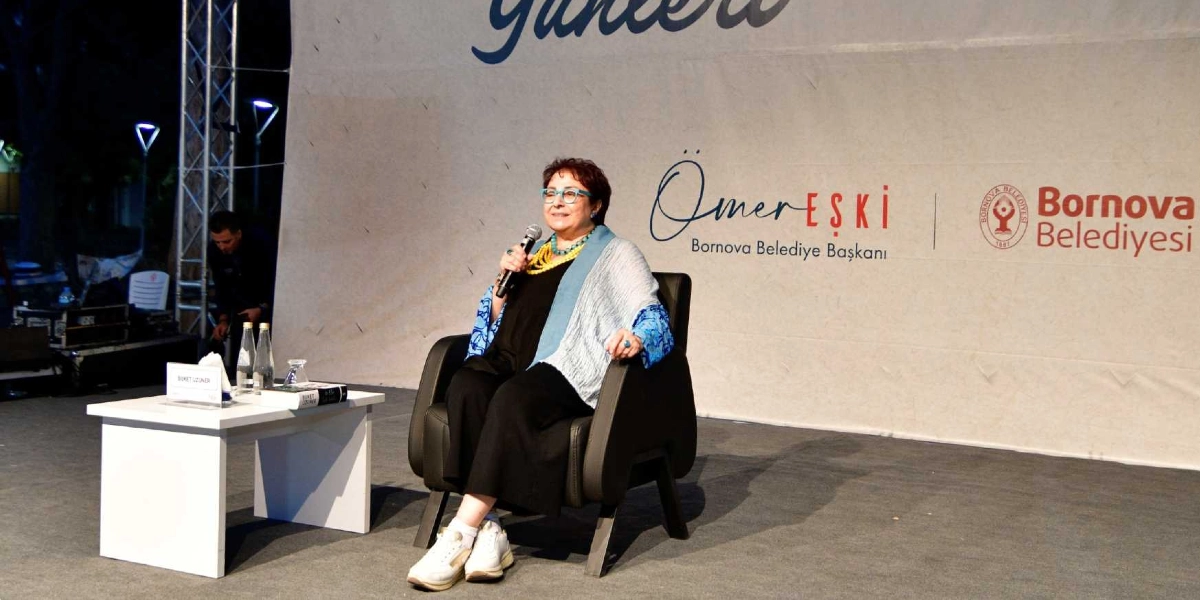 Yazar Buket Uzuner, Bornova Belediyesi tarafından düzenlenen bir etkinlikte sahnede siyah kıyafetleri, mavi desenli şalı, renkli gözlükleri ve spor ayakkabılarıyla siyah bir koltukta oturarak mikrofonla konuşma yapıyor. Yanındaki beyaz masanın üzerinde su şişeleri ve kitapları bulunuyor. Sahne arkasındaki panoda Ömer Eşki Bornova Belediye Başkanı yazısı ve Bornova Belediyesi logosu yer alıyor.
