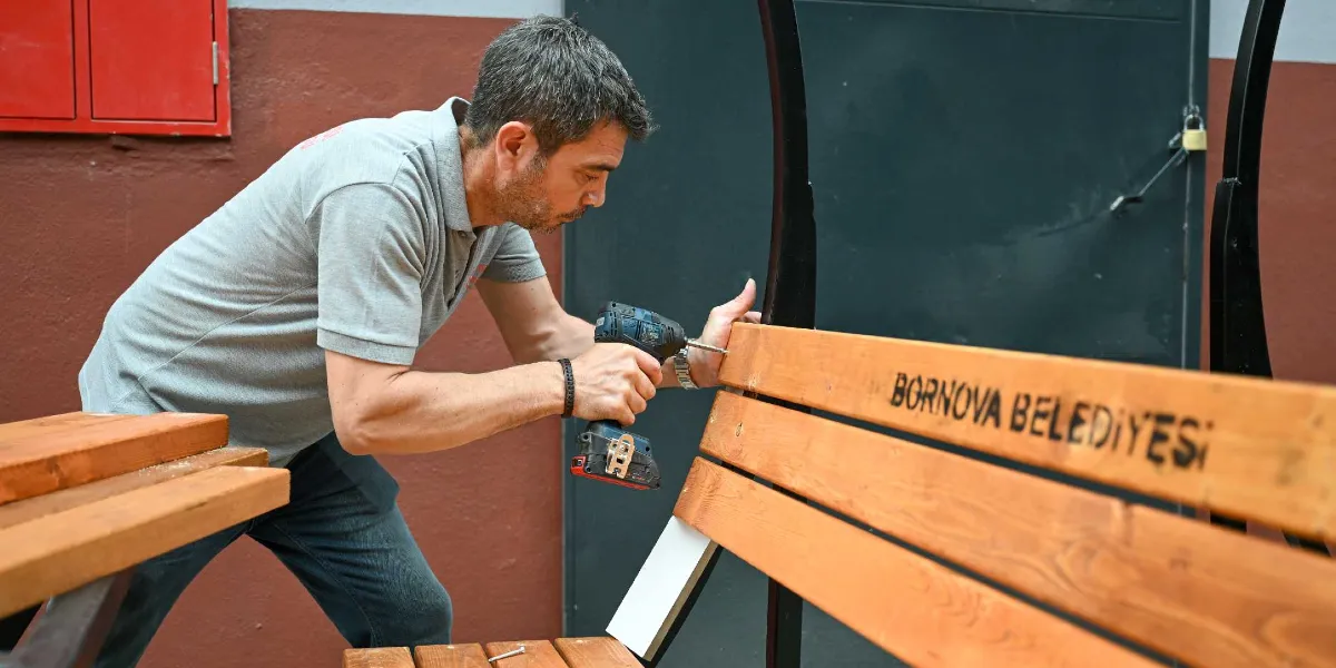 Gri tişörtlü bir belediye çalışanı, üzerinde Bornova Belediyesi yazılı olan ahşap bir park bankının montajını şarjlı vidalama makinesiyle yapıyor.