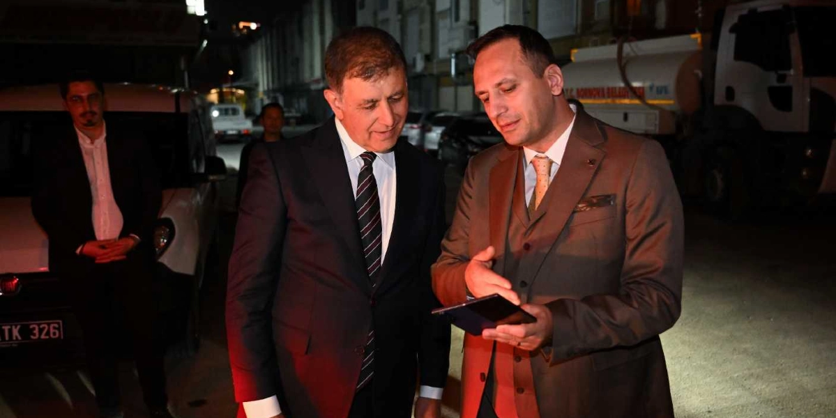 Gece vakti dış mekanda, takım elbiseli iki erkek yan yana durarak ellerindeki tablete bakıyor. Sağdaki kişi tableti tutup bir şeyler anlatırken, soldaki kişi dikkatle onu dinliyor. Arka planda park halindeki araçlar ve Bornova Belediyesi'ne ait bir kamyon görülüyor, sol tarafta ise ellerini önünde birleştirmiş duran başka bir kişi daha yer alıyor.