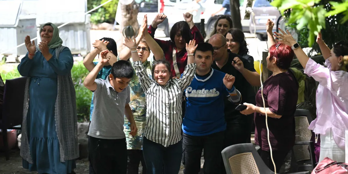 Açık havada bir araya gelen, aralarında özel gereksinimli bireylerin de bulunduğu bir grup insan, ellerini havaya kaldırarak ve gülümseyerek neşeyle kutlama yapıyor. Grubun solunda başörtülü bir kadın alkışlarken, merkezde çizgili gömlekli genç bir kadın ve mavi tişörtlü bir genç erkek ön planda yer alıyor. Etraflarındaki diğer yetişkinler de destekleyici bir tavırla onlara eşlik ediyor.
