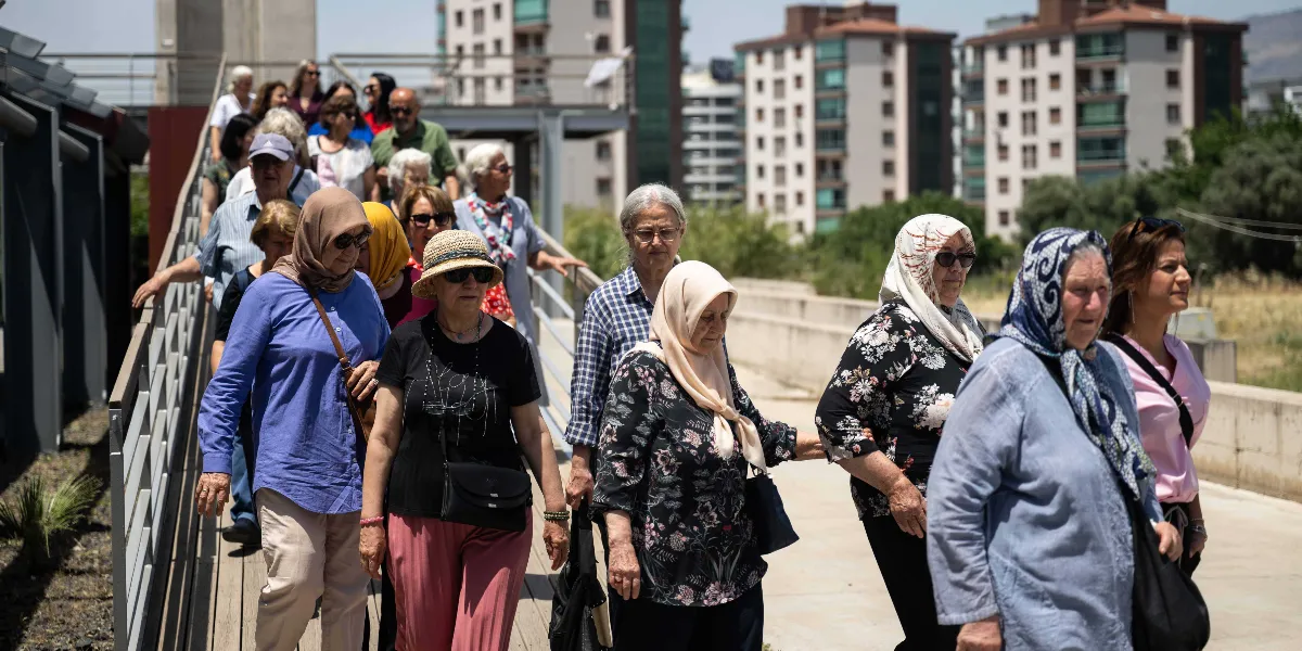 Güneşli bir günde, modern apartmanların bulunduğu bir yerleşkede, ahşap bir yürüyüş yolunda ilerleyen, çoğunluğu orta yaş ve üzeri kadınlardan oluşan kalabalık bir grup. Birçoğu başörtülü olan katılımcılar, yan yana ve peşi sıra yürüyerek sohbet ediyor; bazıları güneş gözlüğü takıyor ve üzerlerinde günlük, rahat kıyafetler bulunuyor.