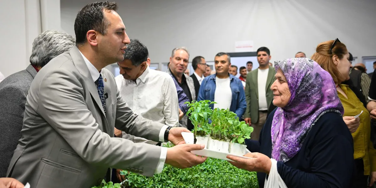 Gri takım elbiseli bir yetkili, mor desenli başörtüsü takan yaşlı bir kadına içinde küçük yeşil fide bulunan bir viyolü uzatıyor. Kapalı bir mekanda gerçekleşen bu etkinlikte, çevredeki diğer insanlar da süreci izliyor.