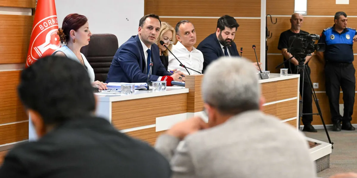 Bornova Belediyesi meclis salonunda, üzerinde Bornova Belediyesi logosunun bulunduğu kırmızı bir bayrağın önünde, bir masa etrafında toplanmış yetkililer toplantı yapmaktadır. Masanın merkezinde konuşma yapan bir erkek yetkili, yanında diğer katılımcılar ve önlerinde mikrofonlar bulunmaktadır. Arka planda bir kameraman ve bir zabıta memuru yer alırken, ön planda toplantıyı izleyen katılımcıların sırtları görünmektedir.