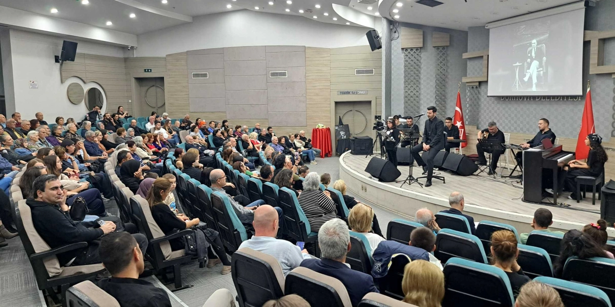 Bornova Belediyesi'ne ait bir konferans salonunda düzenlenen etkinlikte, sahnede mikrofon başında şarkı söyleyen bir çocuk ve ona eşlik eden müzisyenler bulunuyor. Müzisyenler arasında gitar çalan, piyano başında oturan ve şarkı söyleyen yetişkinler yer alıyor. Sahnenin arkasındaki dev ekranda Mustafa Kemal Atatürk'ün siyah beyaz bir fotoğrafı yansıtılıyor ve sahnenin iki yanında Türk bayrakları bulunuyor. Salonu dolduran kalabalık izleyici grubu, sahnedeki performansı dikkatle takip ediyor.