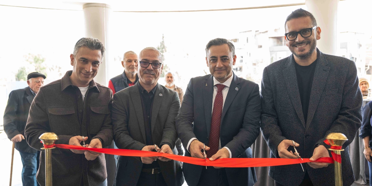 Dört erkek, ellerinde tuttukları makaslarla kırmızı bir açılış kurdelesini kesiyor. Soldan sağa doğru; kahverengi ceketli bir kişi, koyu renk ceketli ve gözlüklü bir kişi, lacivert takım elbiseli ve kırmızı kravatlı bir kişi ve üzerinde koyu renkli bir ceket olan gözlüklü bir kişi yan yana duruyor. Arka planda ise etkinliği izleyen diğer katılımcılar yer alıyor.