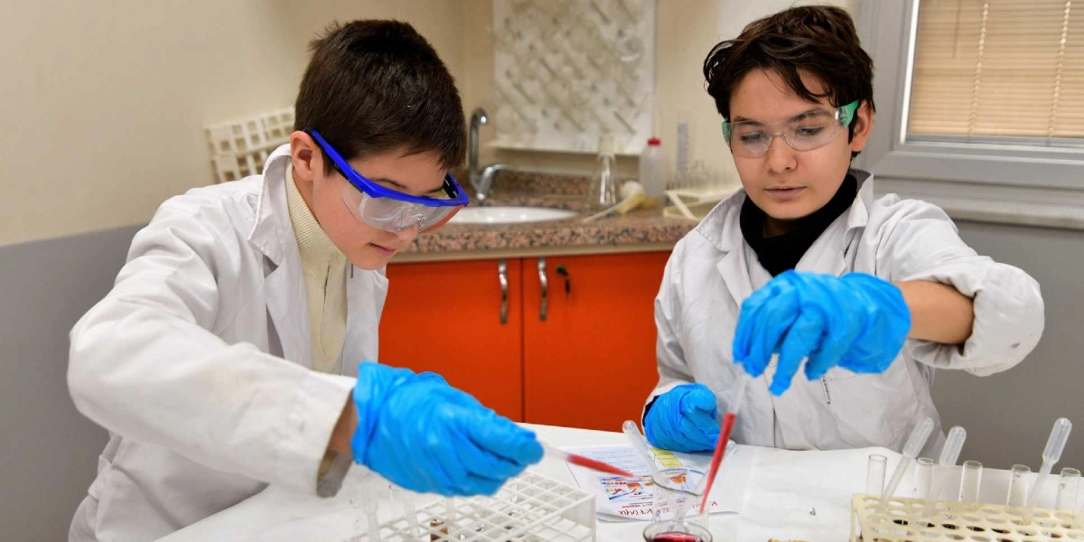 Laboratuvar önlüğü giymiş, koruyucu gözlük ve mavi eldiven takan iki öğrenci, bir masa üzerinde deney tüpleri ve pipetlerle bilimsel çalışma yapıyor. Öğrencilerden biri elindeki pipetle kırmızı renkli bir sıvıyı tüpe aktarırken, diğeri dikkatle çalışmasını sürdürüyor. Arka planda turuncu dolaplı bir laboratuvar tezgahı ve lavabo bulunuyor.