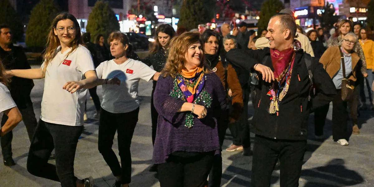 Açık havada, akşam saatlerinde bir araya gelen bir grup insan, yan yana dizilerek halk dansı figürleri sergiliyor. Ön planda, üzerinde Türk bayrağı arması bulunan beyaz tişörtlü kadınlar, mor hırkalı ve renkli fularlı bir kadın ile boynunda kırmızı fular bulunan bir erkek neşeyle dans ediyor. Arka planda ise dansa katılan diğer insanlar ve şehir ışıkları görülüyor.