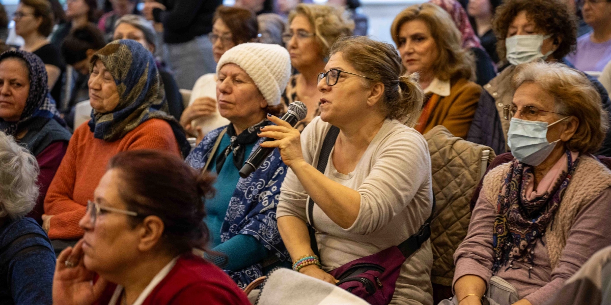 Kapalı bir mekanda düzenlenen etkinlikte, farklı yaş gruplarından kadınlar oturarak dinleyici olarak yer alıyor. Ön planda, gözlüklü bir kadın elinde mikrofon tutarak konuşuyor. Yanındaki kadınlardan bazılarının maske taktığı, bazılarının ise başörtülü olduğu görülüyor. Katılımcılar dikkatle dinliyor ve etkinliğe katılım sağlıyor.