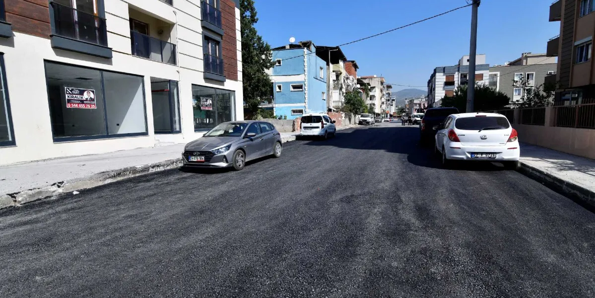Bornova'da yeni asfaltlanmış, pürüzsüz ve temiz bir yolun genel görünümü. Yolun kenarlarında park edilmiş otomobiller, sol tarafta modern bir apartman ve arka planda diğer binalar yer alıyor. Gökyüzü açık ve güneşli bir gün.