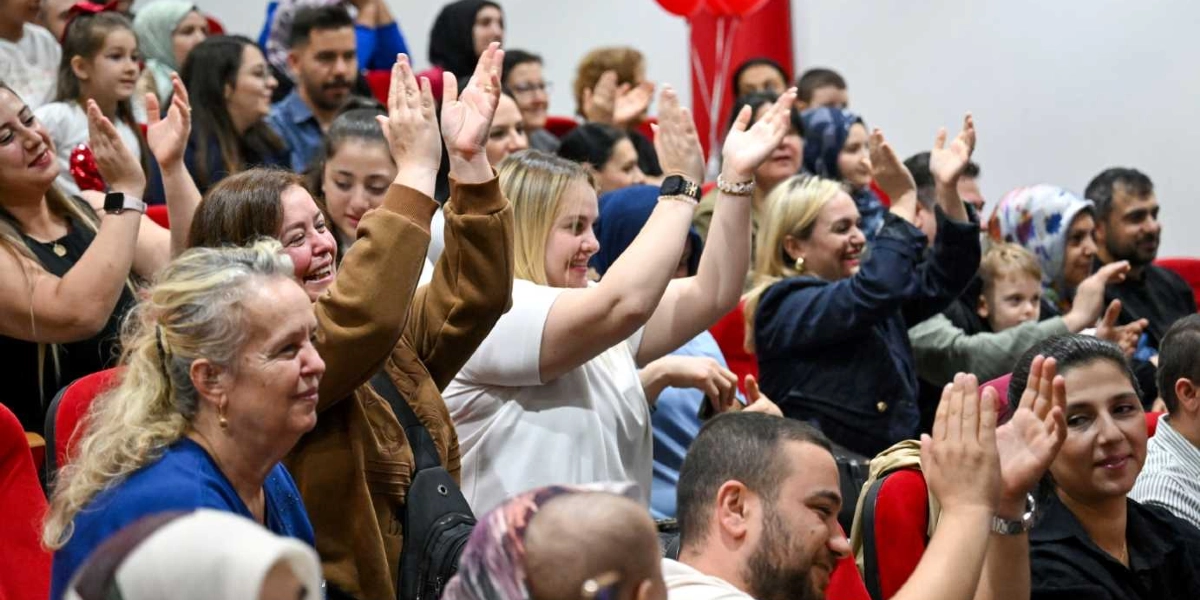 Bir etkinlik salonunda kırmızı koltuklarda oturan ve neşeyle alkışlayan kalabalık bir izleyici grubu. Ön planda, ellerini havaya kaldırarak alkışlayan kadınlar ve gülümseyen yetişkinler yer alırken, arka sıralarda çocuklar ve başörtülü kadınlar da dahil olmak üzere farklı yaş gruplarından insanlar bulunuyor. Ortamda Cumhuriyet coşkusunu yansıtan neşeli ve katılımcı bir atmosfer hakim.