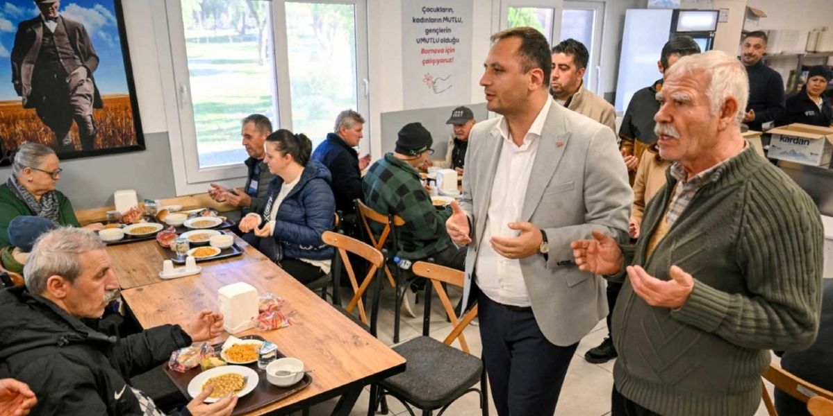 Aydınlık bir yemekhanede, masalarda oturan vatandaşların ve ayakta duran bir yetkilinin ellerini açarak dua ettiği bir an. Masalarda yemek tepsileri ve tabaklar bulunurken, duvarda Mustafa Kemal Atatürk'ün bir portresi ve üzerinde 