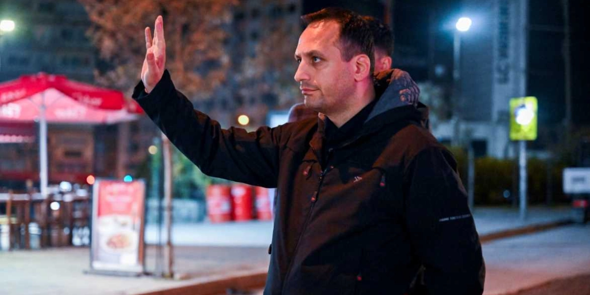 Gece vakti dışarıda duran, siyah montlu bir erkek, sağ elini havaya kaldırarak birini veya bir şeyi durdurmaya çalışıyor. Arka planda ise bulanık bir şekilde dış mekanda bulunan masa ve sandalyeler ile kırmızı renkli variller görülüyor.
