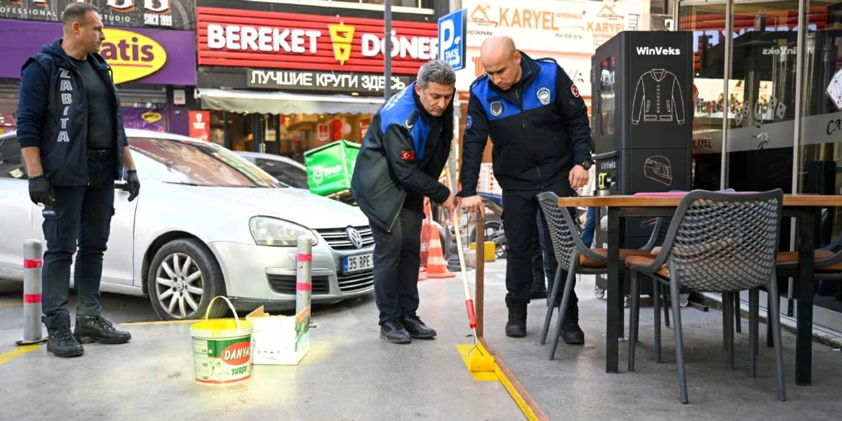 Bornova'da zabıta ekipleri, bir işletmenin önündeki kaldırımda yer alan sınırları belirlemek için sarı boya ile çizgi çekiyor. Üniformalı üç zabıta memurundan biri elindeki rulo fırçayla yeri boyarken, diğer ikisi çalışmayı takip ediyor. Arka planda 