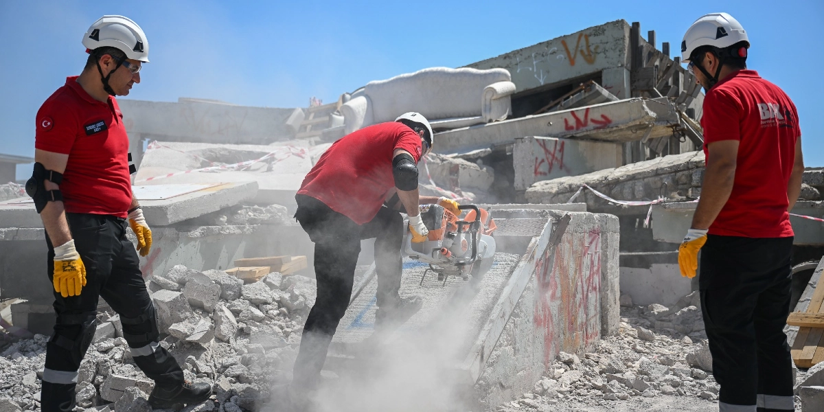 Beyaz baretli, kırmızı tişörtlü ve iş eldivenli üç arama kurtarma görevlisi, yıkılmış bir binanın enkazında çalışıyor. Ortadaki görevli, beton bir bloğu kesmek için motorlu beton kesme makinesi kullanırken etrafa yoğun toz yayılıyor. Diğer iki görevli ise çevreyi kontrol ederek çalışmaya eşlik ediyor. Arka planda enkazın üzerinde kalmış bir koltuk ve yıkıntıların arasında güvenlik şeritleri görülüyor.
