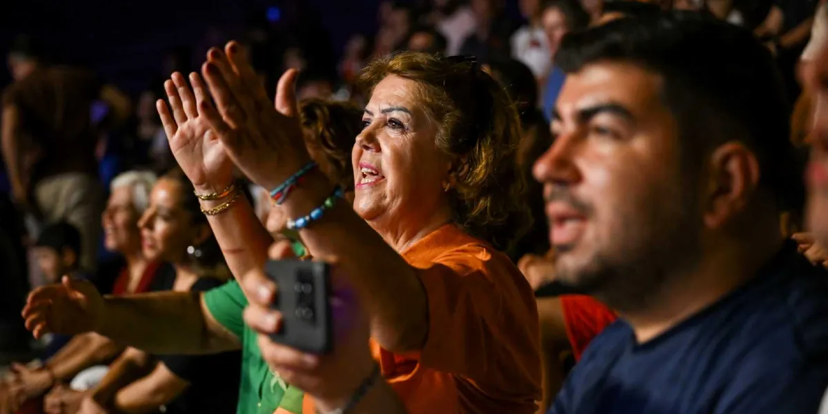 Karanlık bir ortamda düzenlenen bir etkinlikte, turuncu tişörtlü orta yaşlı bir kadın coşkuyla ellerini havaya kaldırıp alkışlarken, yanında koyu renk tişörtlü bir erkek izleyici heyecanla eşlik ediyor. Arka planda kalabalık bir izleyici kitlesi seçiliyor.