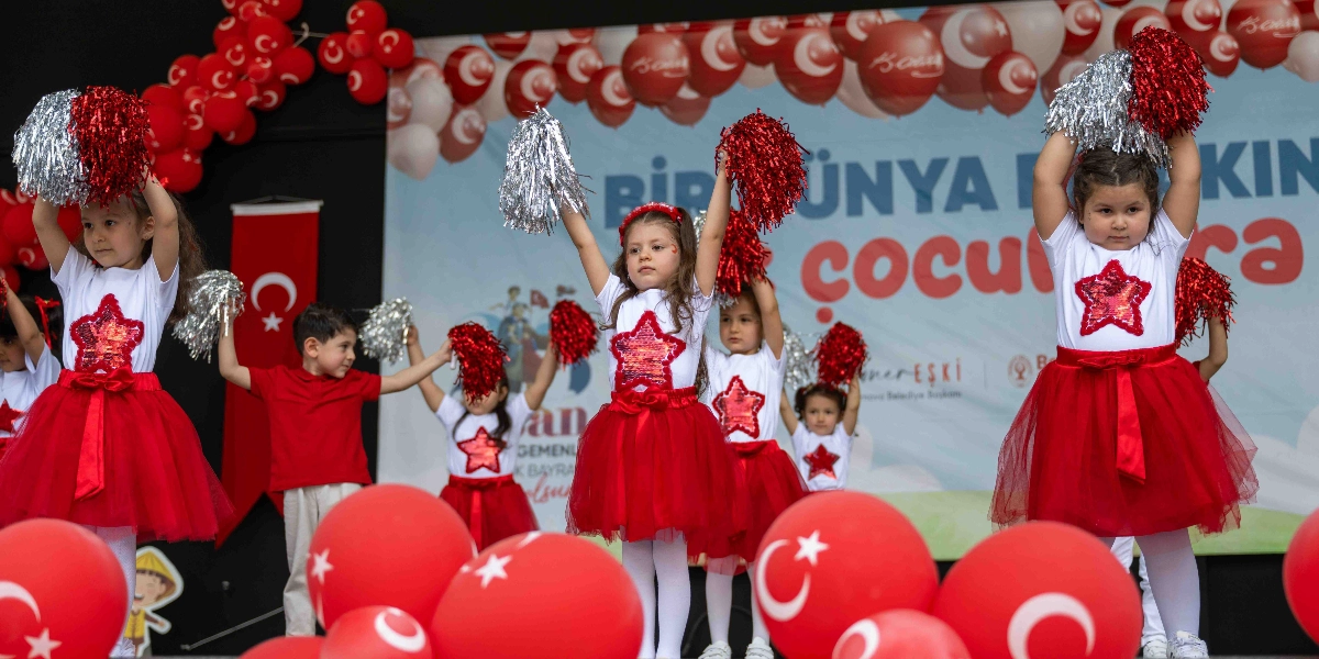 23 Nisan Ulusal Egemenlik ve Çocuk Bayramı kutlamaları kapsamında sahnede dans eden, üzerinde kırmızı yıldız işlemeli beyaz tişört ve kırmızı tül etek bulunan çocuklar. Çocukların bir kısmı ellerinde kırmızı ve gümüş renkli ponponlar tutuyor. Sahnenin arka planında üzerinde 