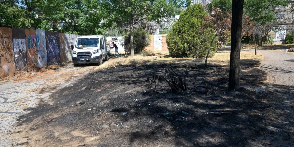 Bornova Belediyesi'ne ait beyaz bir kamyonetin park halinde olduğu, grafiti yazılarıyla kaplı metal panellerle çevrili, bir kısmı yanmış ve kararmış toprak alanın temizlik çalışmaları için düzenlendiği açık hava alanı.