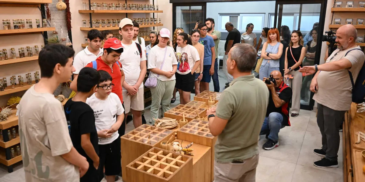 Bornova'da bir tohum merkezinde, ahşap raflarda dizili cam kavanozların bulunduğu bir ortamda, rehberlik yapan bir kişinin etrafında toplanmış gençlerden oluşan bir grup. Rehber elini uzatarak açıklamalarda bulunurken, gruptakiler ilgiyle onu dinliyor, bir fotoğrafçı ise bu anı ölümsüzleştirmek için çömelmiş çekim yapıyor. Ortadaki ahşap tezgahlarda tohum örnekleri ve küçük havanlar yer alıyor.