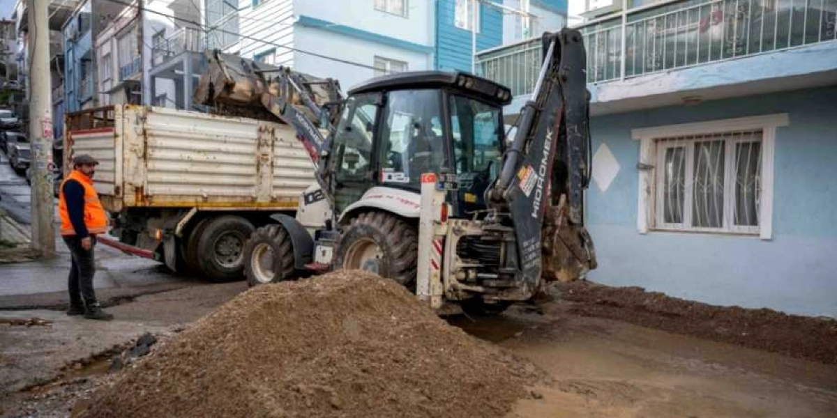 Bornova'da bir sokakta sel sonrası temizlik çalışması yapan bir iş makinesi ve yanında duran turuncu yelekli bir görevli. Hidromek marka kepçe, yoldaki çamur ve toprak yığınlarını bir kamyonun kasasına yüklüyor.