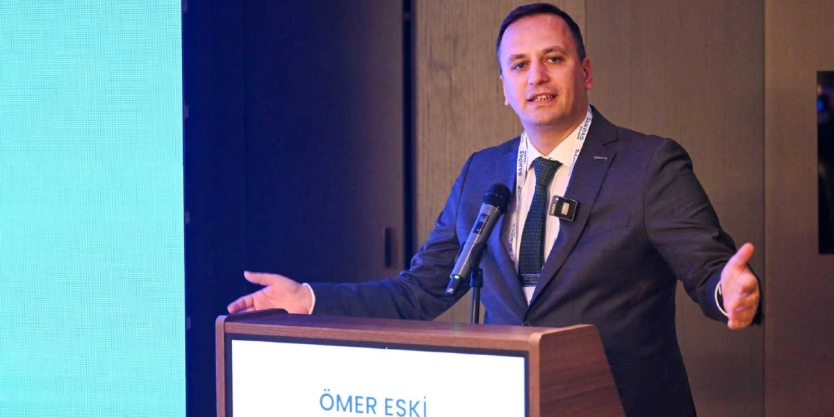 Koyu renk takım elbiseli ve yeşil kravatlı Ömer Eşki, bir kürsü arkasında konuşma yapıyor. Eşki, ellerini iki yana açarak hitap ettiği sırada boynunda üzerinde 