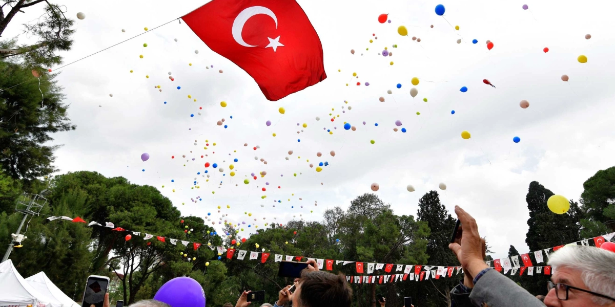 Açık havada düzenlenen bir etkinlikte, gökyüzüne bırakılmış çok sayıda renkli balon ve dalgalanan büyük bir Türk bayrağı görülüyor. Kalabalık bir grup insan, ellerindeki telefonlarla bu anı kaydediyor. Arka planda ağaçlar ve Türk bayraklarıyla süslenmiş bir alan yer alıyor.