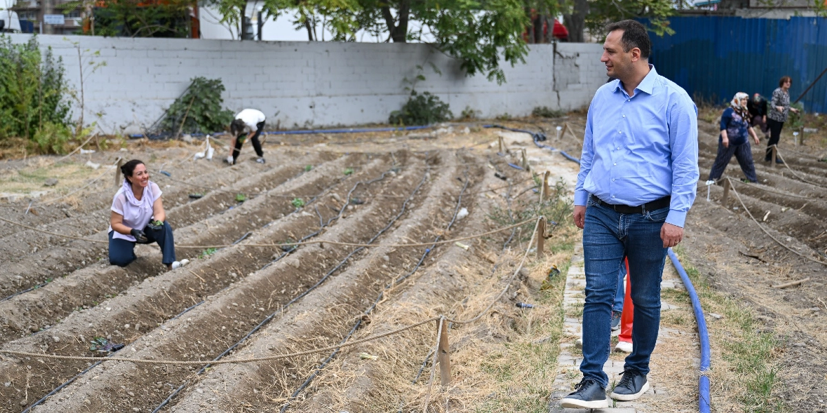 Mavi gömlekli ve kot pantolonlu bir erkek, Bornova'da kadınların toprakla uğraştığı, sıralı ekim alanlarının bulunduğu bir bahçede yürürken gülümsüyor. Arka planda toprakla ilgilenen ve fide diken kadınlar görülüyor.