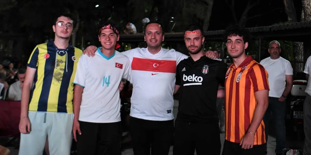 Gece vakti dış mekanda yan yana duran, farklı Türk futbol takımlarının formalarını giymiş beş genç erkek. Soldan sağa doğru; sarı-lacivert çubuklu Fenerbahçe forması giyen gözlüklü bir genç, üzerinde 14 numaralı beyaz Türkiye milli takım forması olan bir genç, ortada beyaz üzerine kırmızı şeritli Türkiye milli takım forması giyen bir adam, siyah Beşiktaş forması giyen bir genç ve sarı-kırmızı çubuklu Galatasaray forması giyen bir genç yer alıyor. Arka planda ise bulanık şekilde diğer insanlar ve bir motosiklet görülüyor.