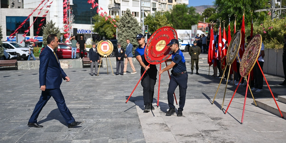 Bornova'da düzenlenen 19 Eylül Gaziler Günü töreninde, iki zabıta memuru üzerinde Bornova Belediyesi amblemi bulunan kırmızı renkli çelengi Atatürk Anıtı'na yerleştirmek üzere taşırken, lacivert takım elbiseli bir yetkili tören alanında yürümektedir. Arka planda Türk bayrakları, törene katılan gaziler ve diğer yetkililer yer almaktadır.