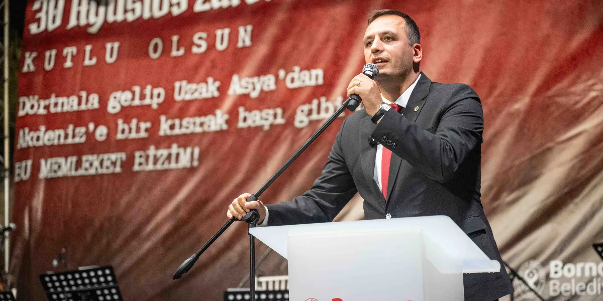 Koyu renk takım elbise ve kırmızı kravat giymiş bir erkek, üzerinde Bornova Belediyesi logosu bulunan beyaz bir kürsüde mikrofonla konuşma yapıyor. Arkasında, üzerinde 